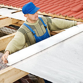 Ein Arbeiter zieht eine flammhemmende Unterdachfolie über Dachbalken eines halb gedeckten Dachtes | Abifor